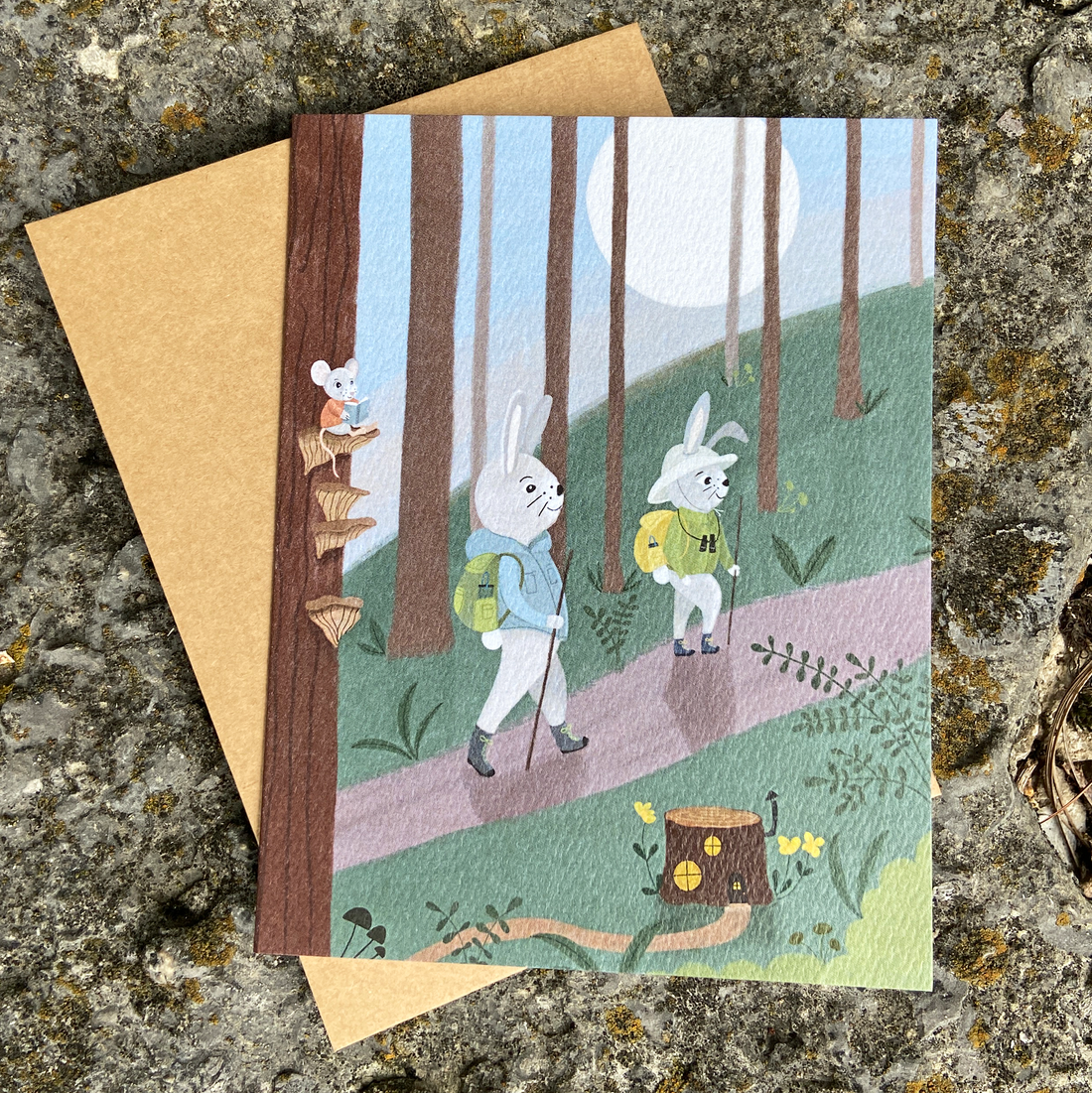 Illustrated greeting card featuring two rabbits hiking in the woods with a mouse reading on mushroom steps