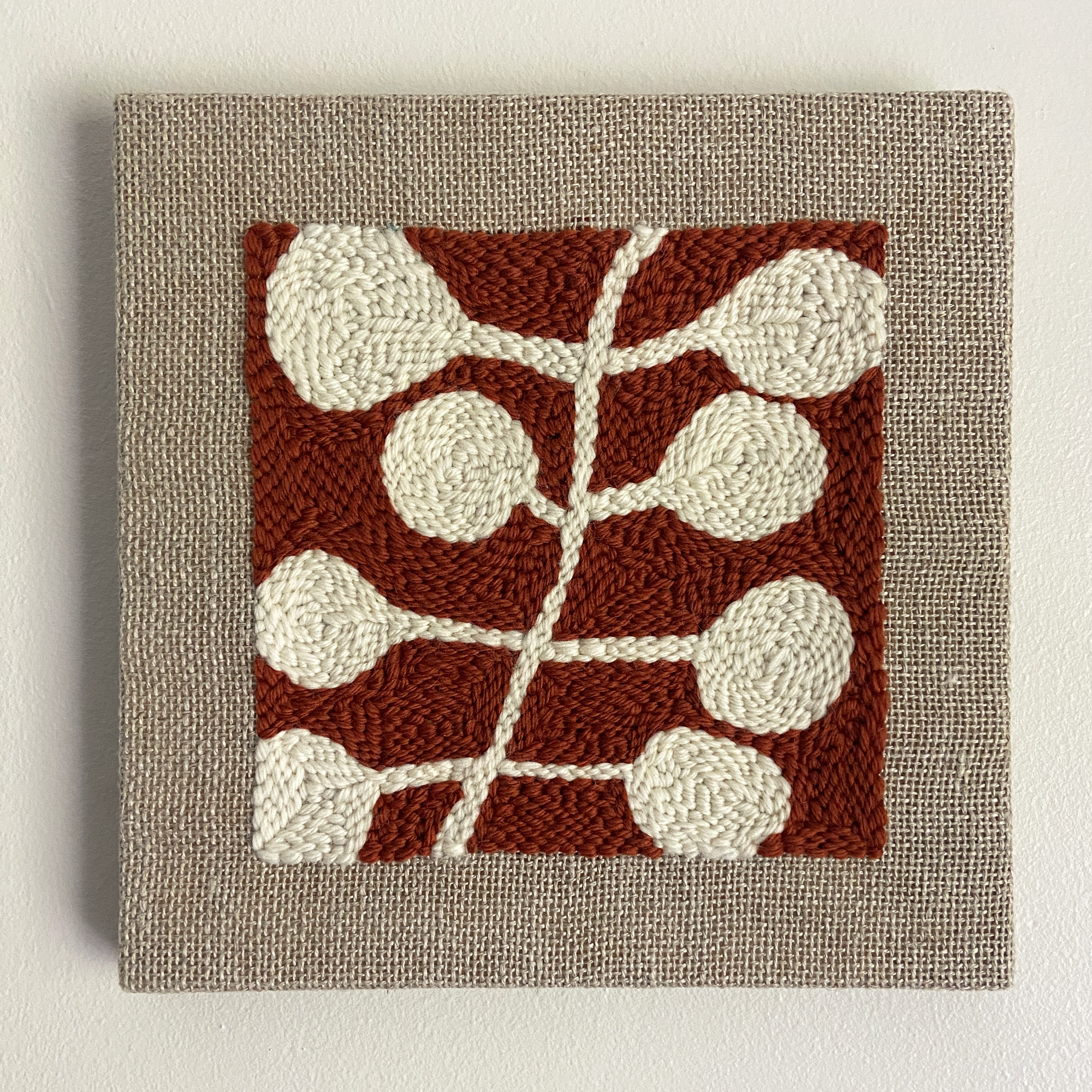 A square woven wall hanging or tapestry with a textured beige linen border and a central rust red square featuring an abstract cream leaf or vine pattern.