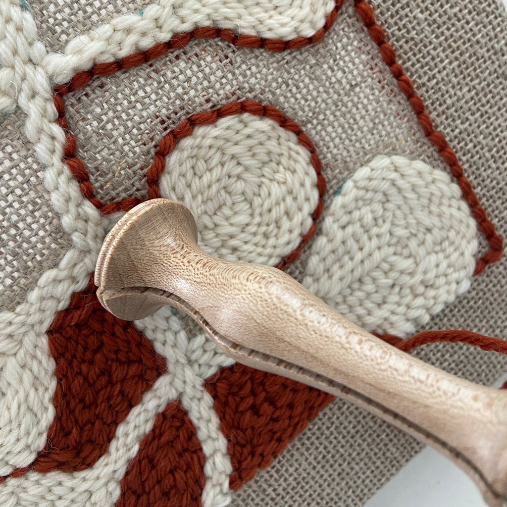 Close-up of a textured needlepoint canvas with cream and rust-brown raised shapes on a beige mesh background, with a wooden-handled punch needle tool resting across the surface.