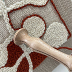 Close-up of a textured needlepoint canvas with cream and rust-brown raised shapes on a beige mesh background, with a wooden-handled punch needle tool resting across the surface.