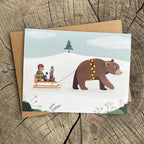 Card with illustration of a child and cat on a sled pulled by a bear, on a wooden surface