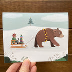 Illustrated card with a bear pulling a sled with a child, cat and stack of gifts