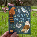 Happy birthday birds card with bird illustrations and 'Happy Birtdhday' text held in front of a natural background with green grass and pink flowers.