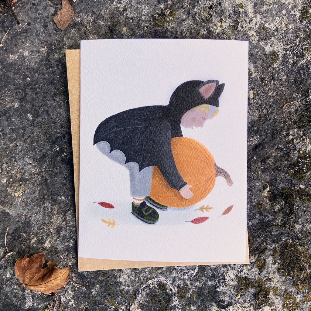 Fall greeting card with a child in a bat costume picking up a pumpkin, placed on a textured stone surface.