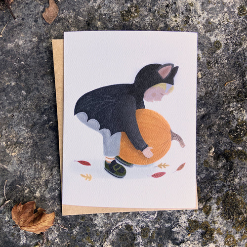 Fall greeting card with a child in a bat costume picking up a pumpkin, placed on a textured stone surface.