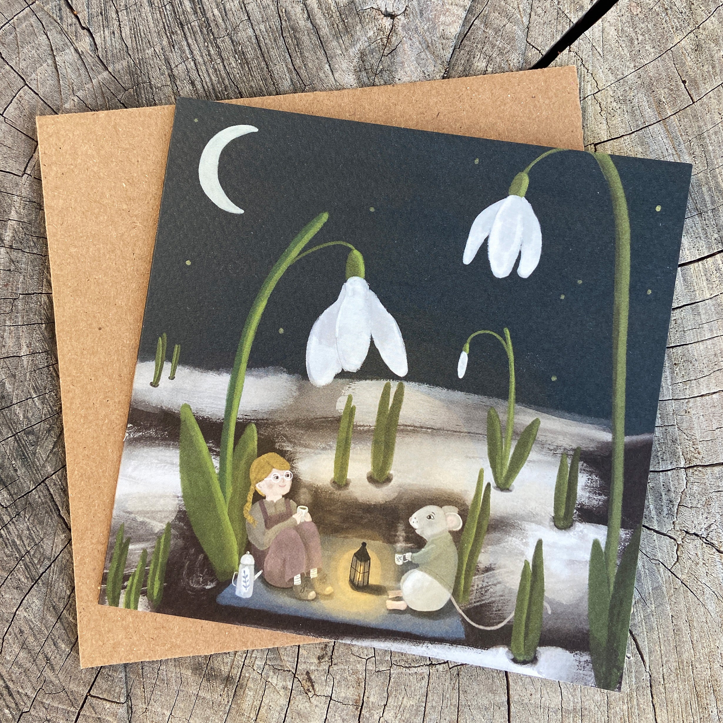 Greeting card with illustration of a girl and mouse drinking tea in a snowdrop garden, on a wooden surface