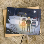 Christmas greeting card with illustration of a child and two cats caroling in a snowy scene, placed on a wooden surface.