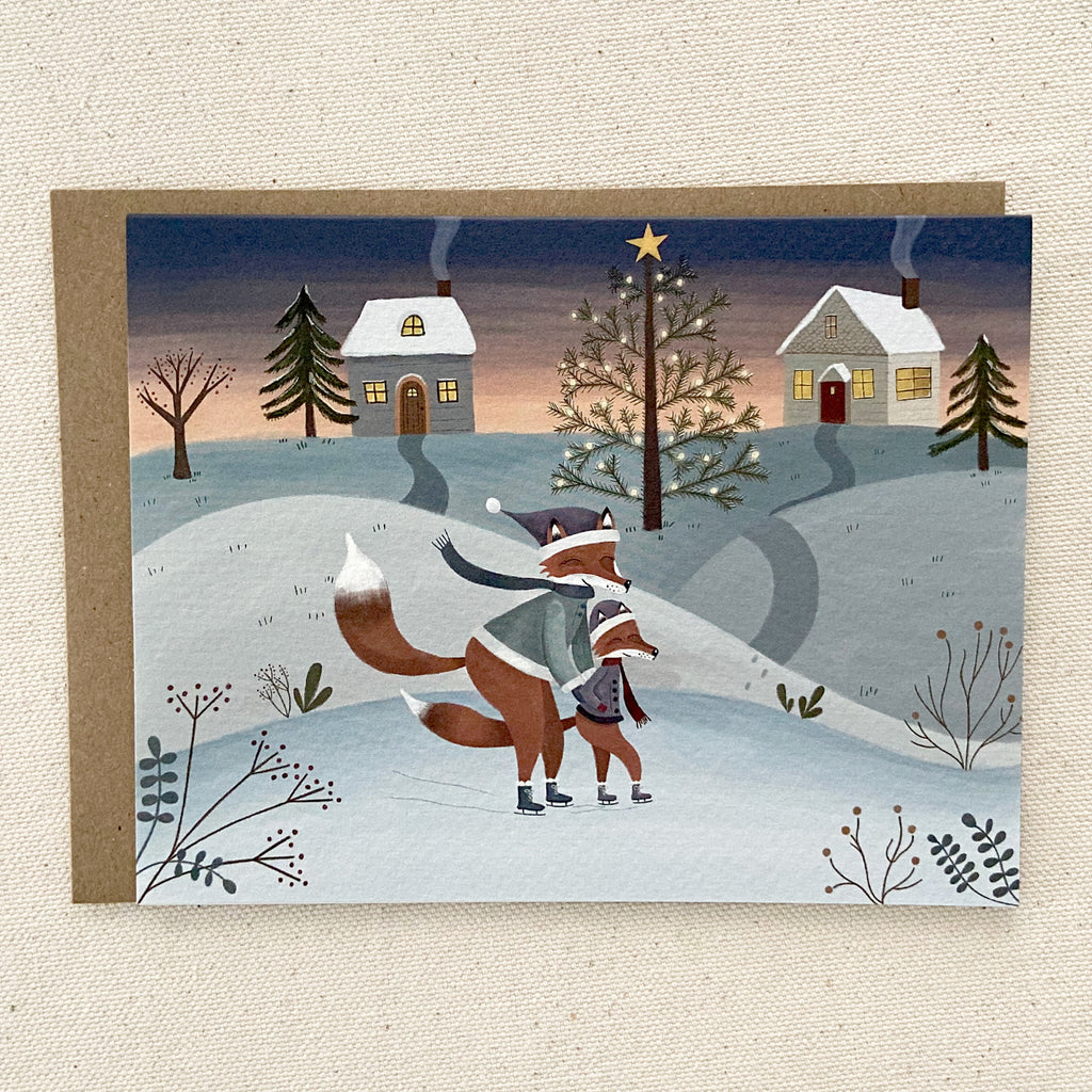 Greeting card with winter scene featuring two foxes, a house, and a Christmas tree.