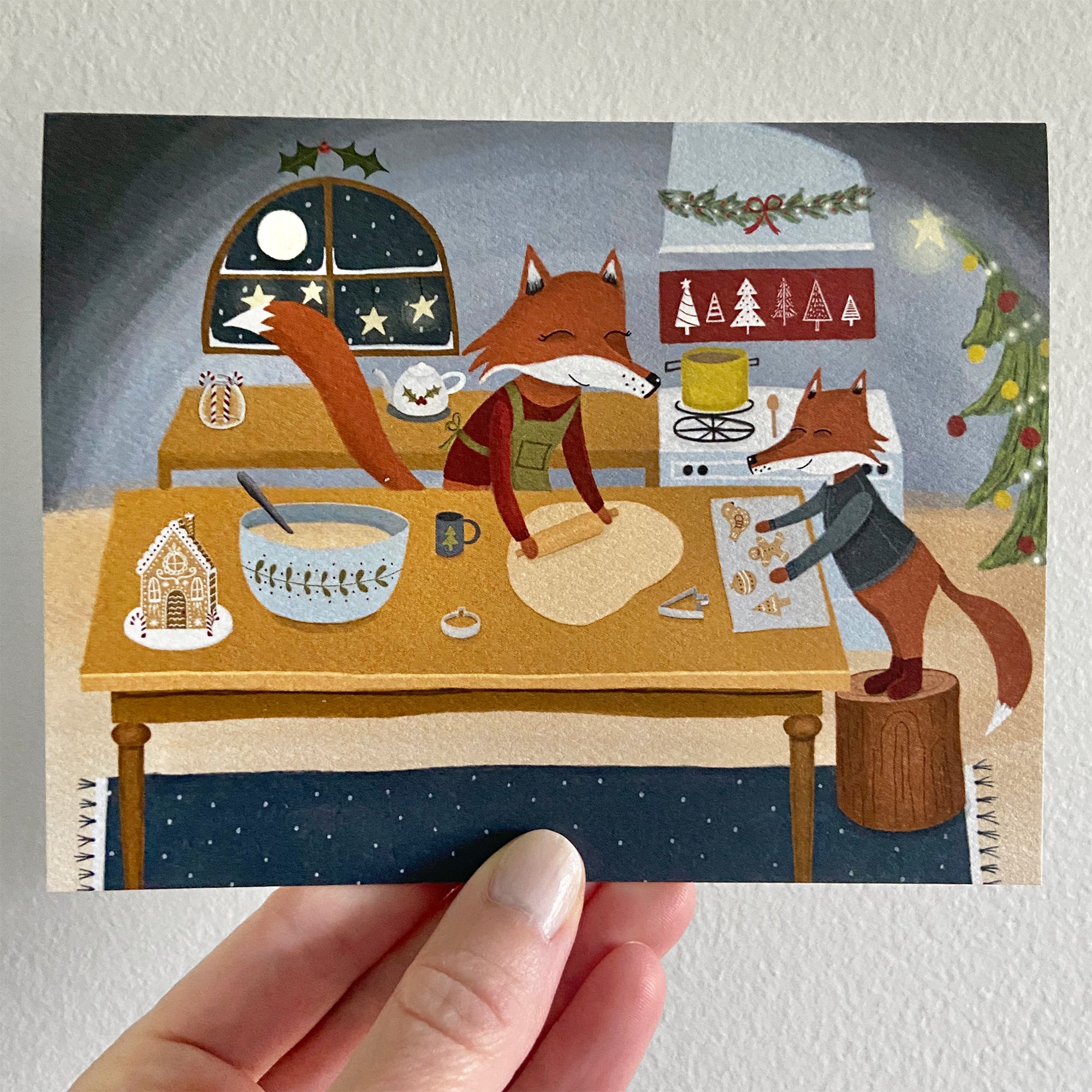 Illustrated Christmas card of two foxes baking gingerbread cookies in a cozy kitchen.
