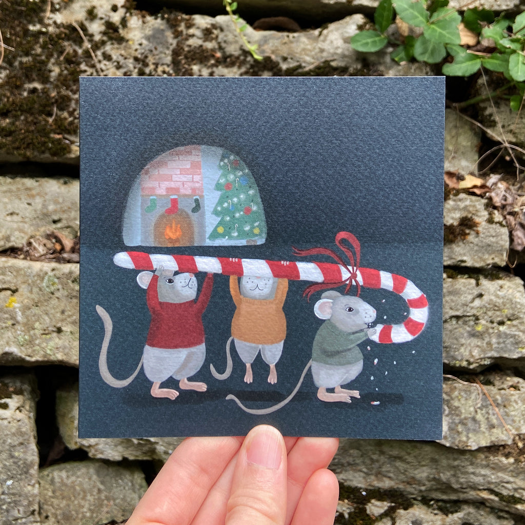 Greeting card of three mice holding a candy cane with a fireplace and Christmas tree in the background, held against a stone wall.