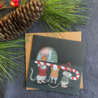 Christmas card with mice holding a candy cane, surrounded by pine cones and branches on a gray fabric background.