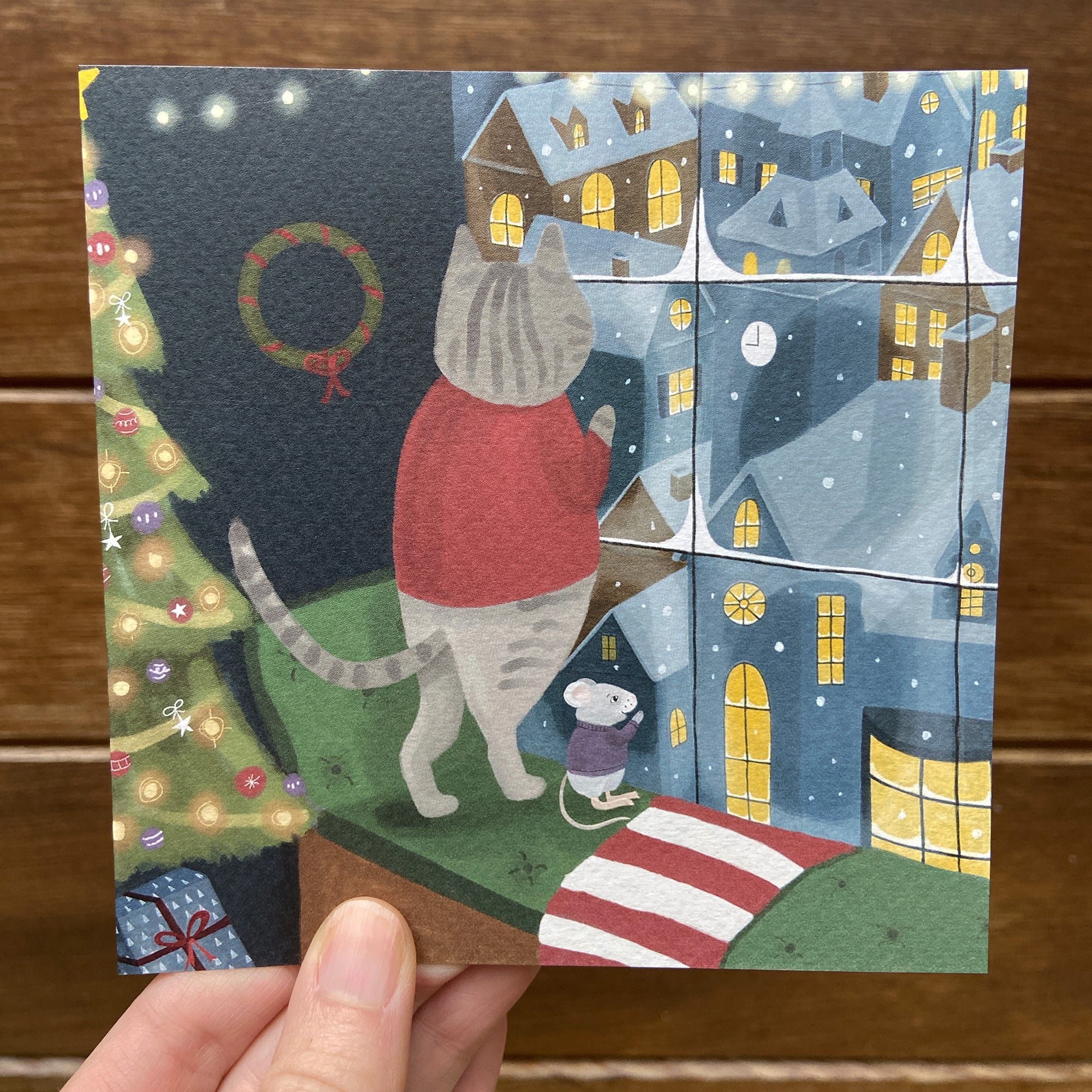Hand holding a Christmas-themed card with a cat and mouse looking out the window by a Christmas tree.