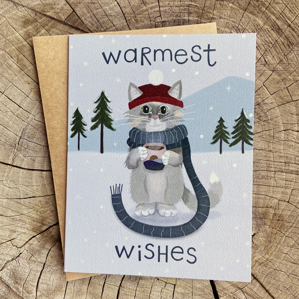 Winter greeting card featuring a cat sipping hot chocolate while wearing a scarf in a snowy setting with hand lettered type saying "Warmest Wishes"