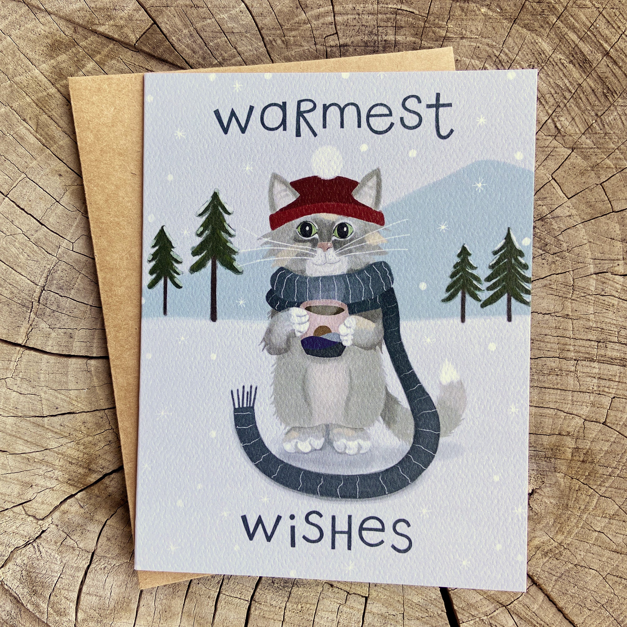 Winter greeting card featuring a cat sipping hot chocolate while wearing a scarf in a snowy setting with hand lettered type saying "Warmest Wishes"