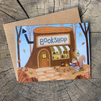 An illustrated card showing a whimsical woodland bookshop built into a tree stump, with a squirrel reading outside and books in the window, placed on top of a kraft envelope over a wood-grain surface.