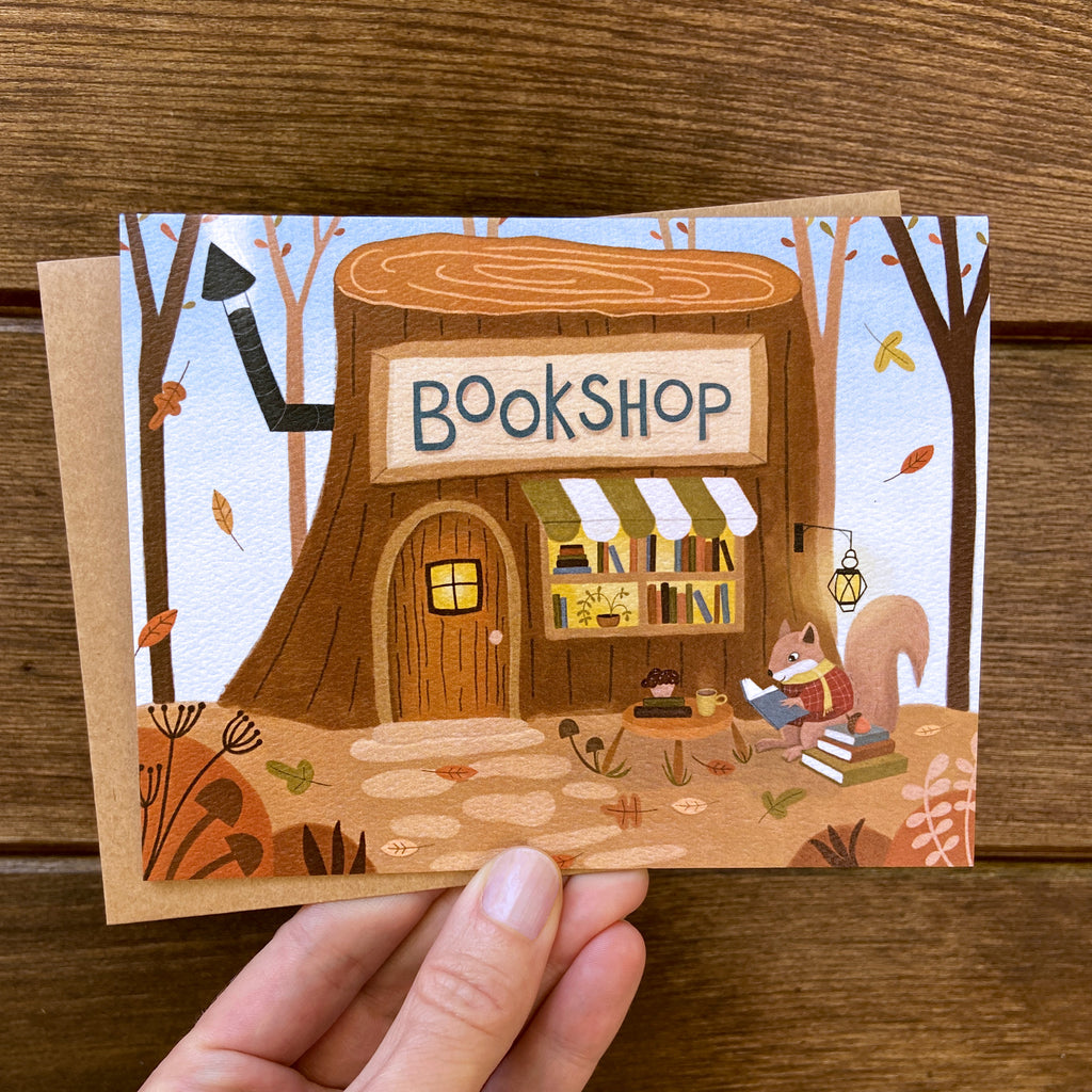 A hand holds an illustrated card featuring a whimsical bookshop built into a tree stump, with autumn trees, leaves, a squirrel reading, and the words “BOOKSHOP” on the front. The card is photographed against a wooden surface.