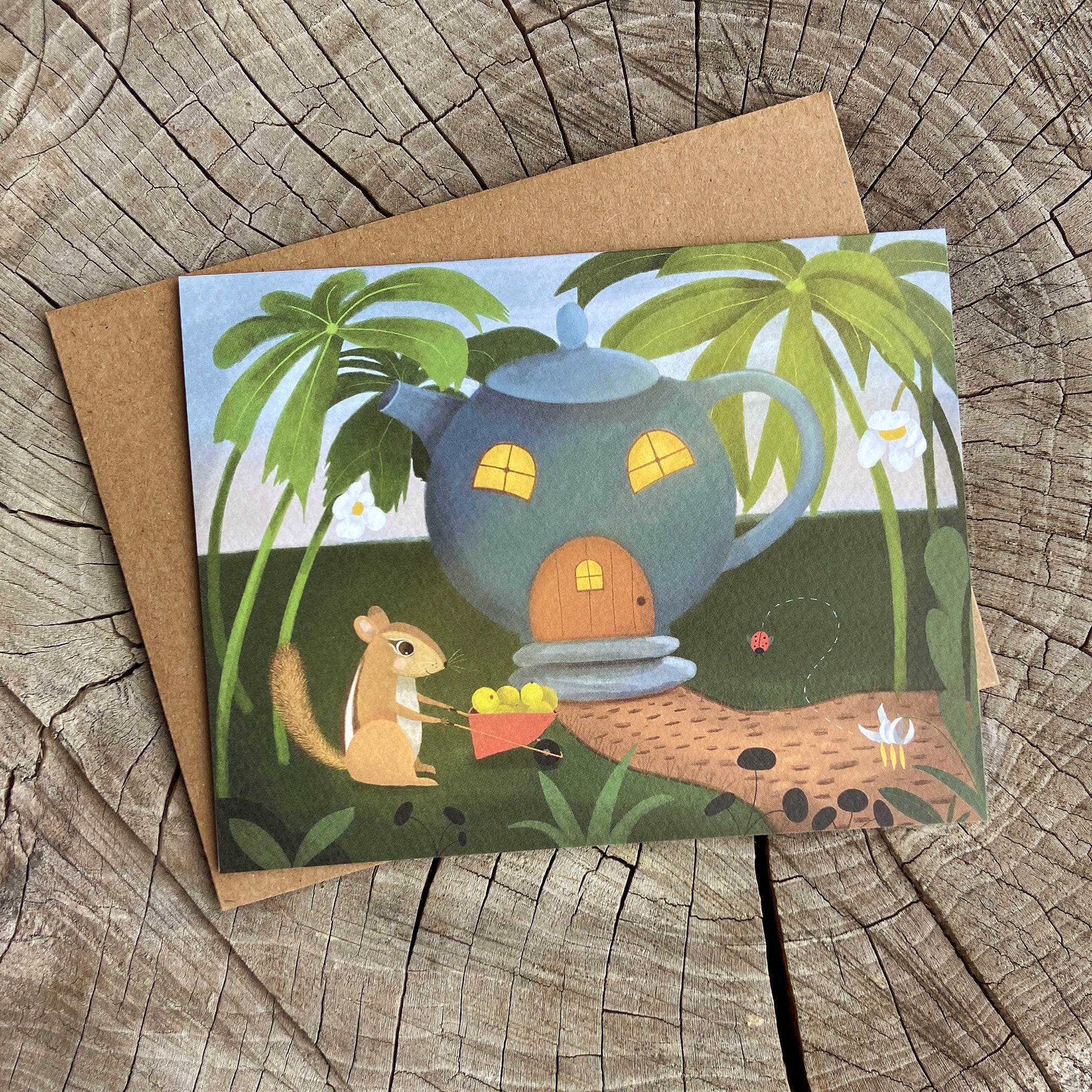 Greeting card with a teapot house and chipmunk illustration on a wooden surface