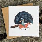 Greeting card with illustration of a child riding a fox, set against a dark blue night sky with stars and trees. The card is placed on a textured stone surface.