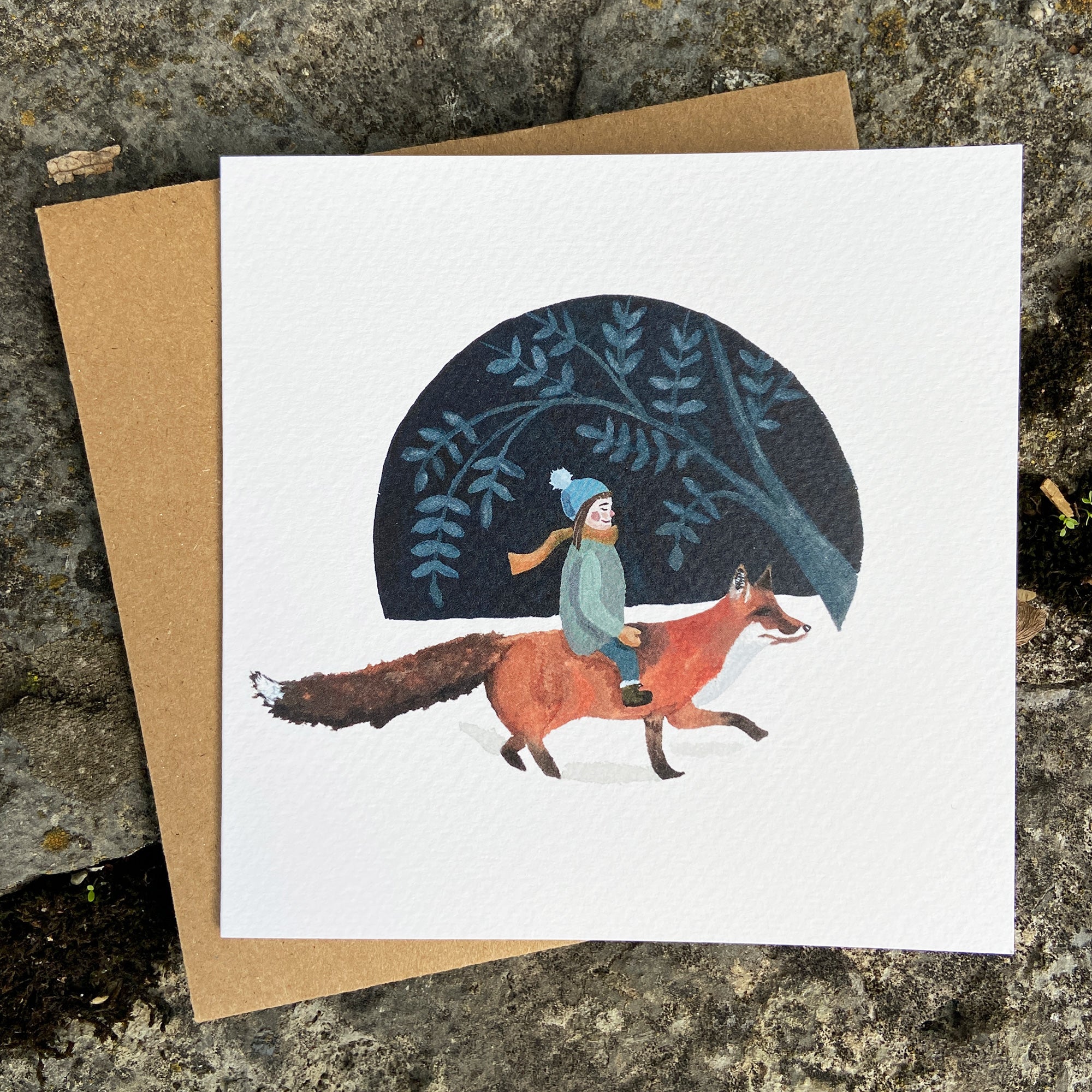 Greeting card with illustration of a child riding a fox, set against a dark blue night sky with stars and trees. The card is placed on a textured stone surface.