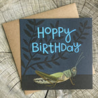 Birthday card with grasshopper illustration and 'Hoppy Birthday' text, on a wooden surface