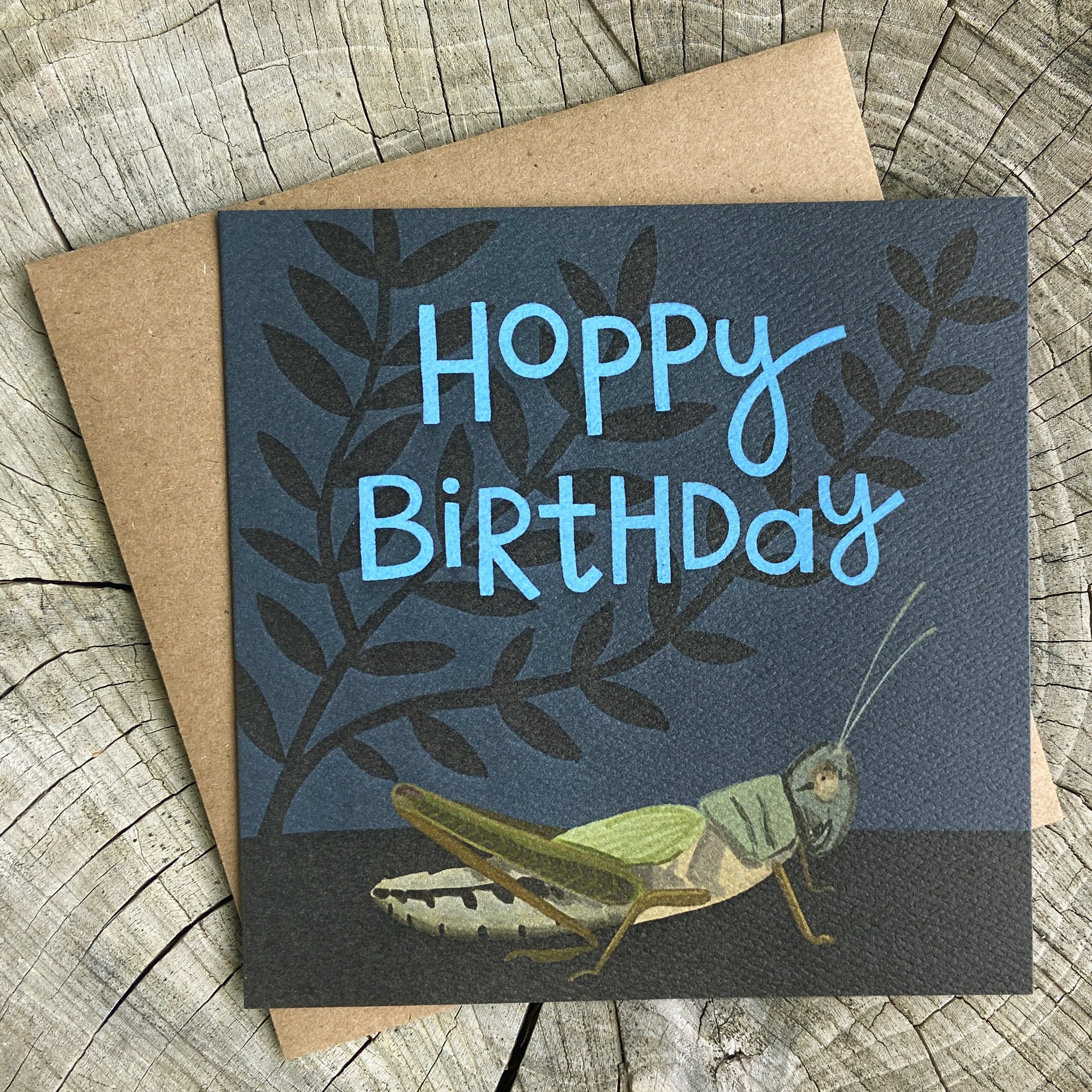 Birthday card with grasshopper illustration and 'Hoppy Birthday' text, on a wooden surface