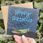 Birthday card with grasshopper illustration and 'Hoppy Birthday' text, held against a natural background.