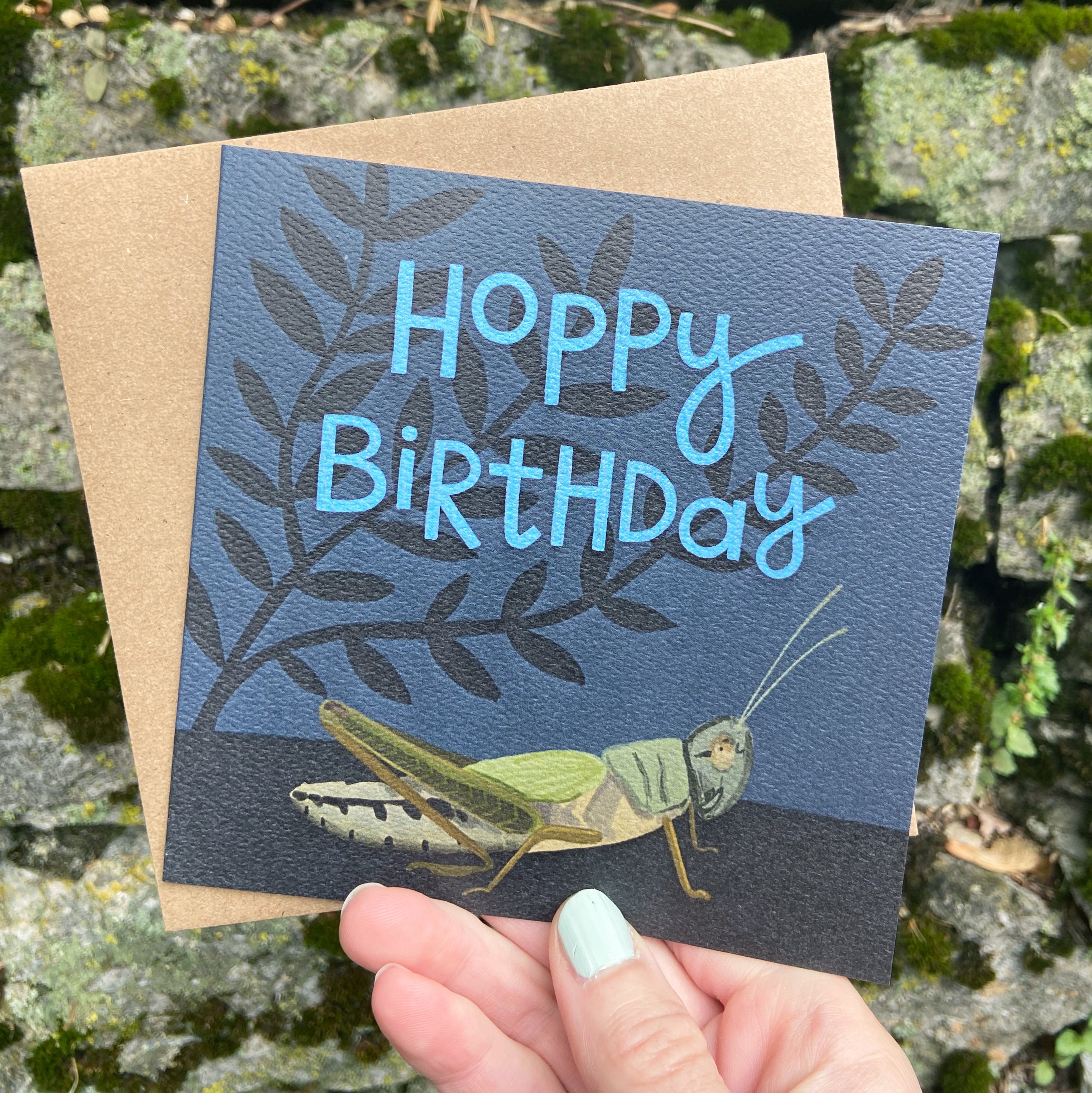 Birthday card with grasshopper illustration and 'Hoppy Birthday' text, held against a natural background.