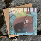 Card with illustration of two bears hugging in a forest setting on a textured stone surface.