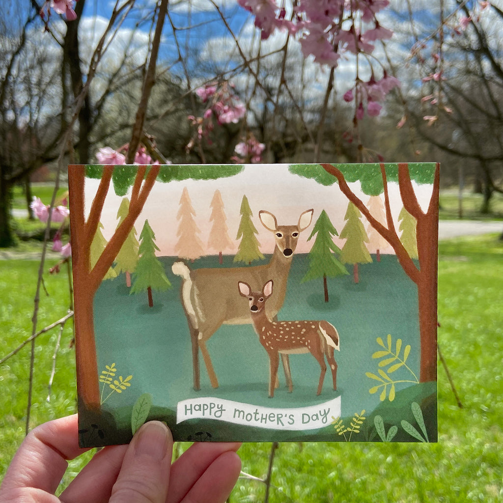 Card with deer illustration and 'Happy Mother's Day' text held in front of a natural background with trees and flowers.