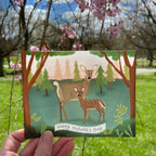 Card with deer illustration and 'Happy Mother's Day' text held in front of a natural background with trees and flowers.