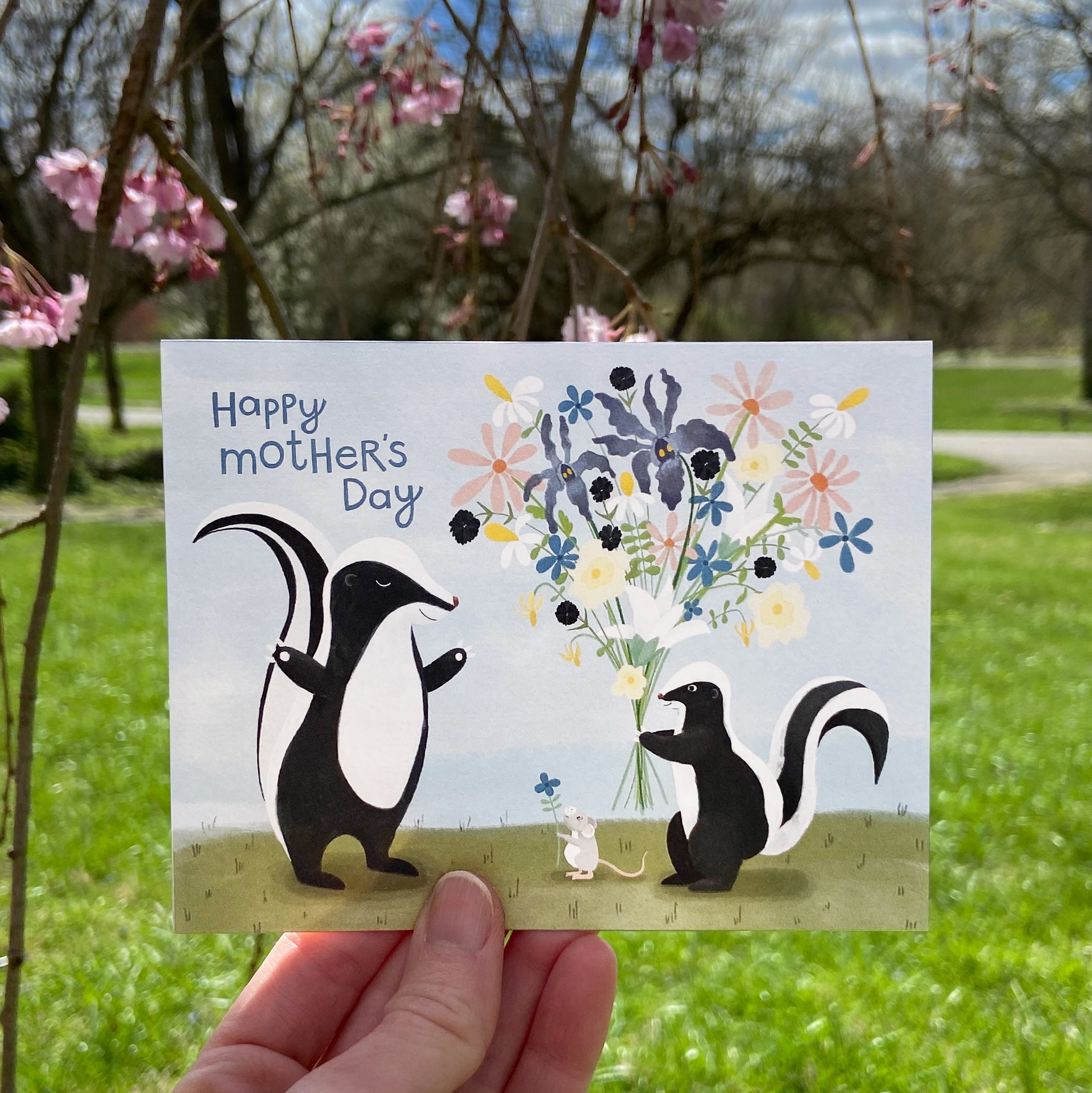 Hand holding a Mother's Day card with skunks and flowers in a park setting