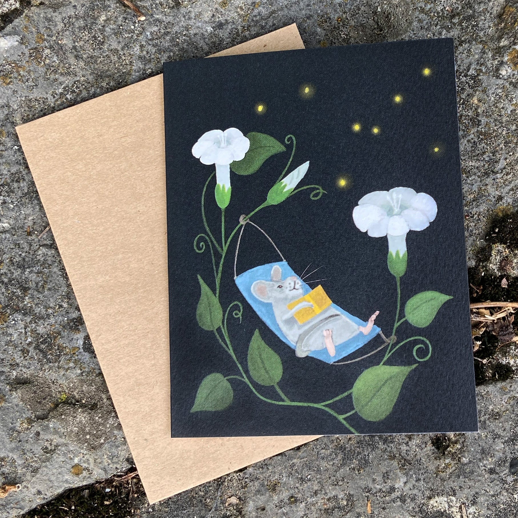 Greeting card with a mouse reading a book among flowers on a dark background, placed on a textured surface.
