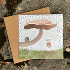 Woodland greeting card with a mushroom illustration and a mouse character, placed on a textured surface.