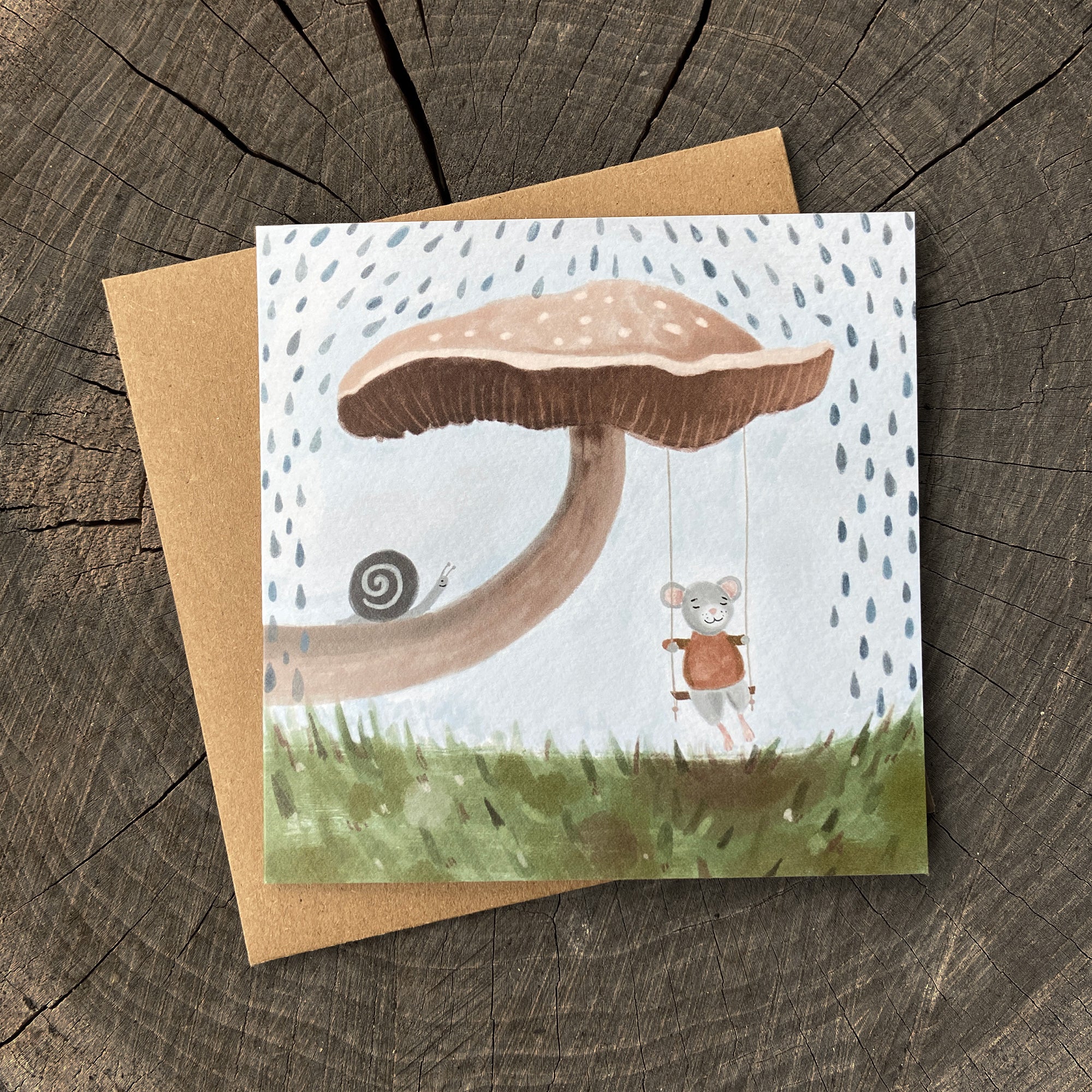 Greeting card with a mouse swinging from a mushroom with a snail nearby, on a wooden surface