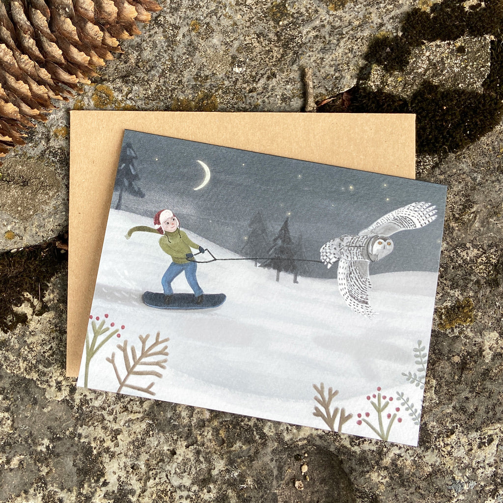 Greeting card with a snowy scene of a boy snowboarding and an owl, on a textured stone surface.