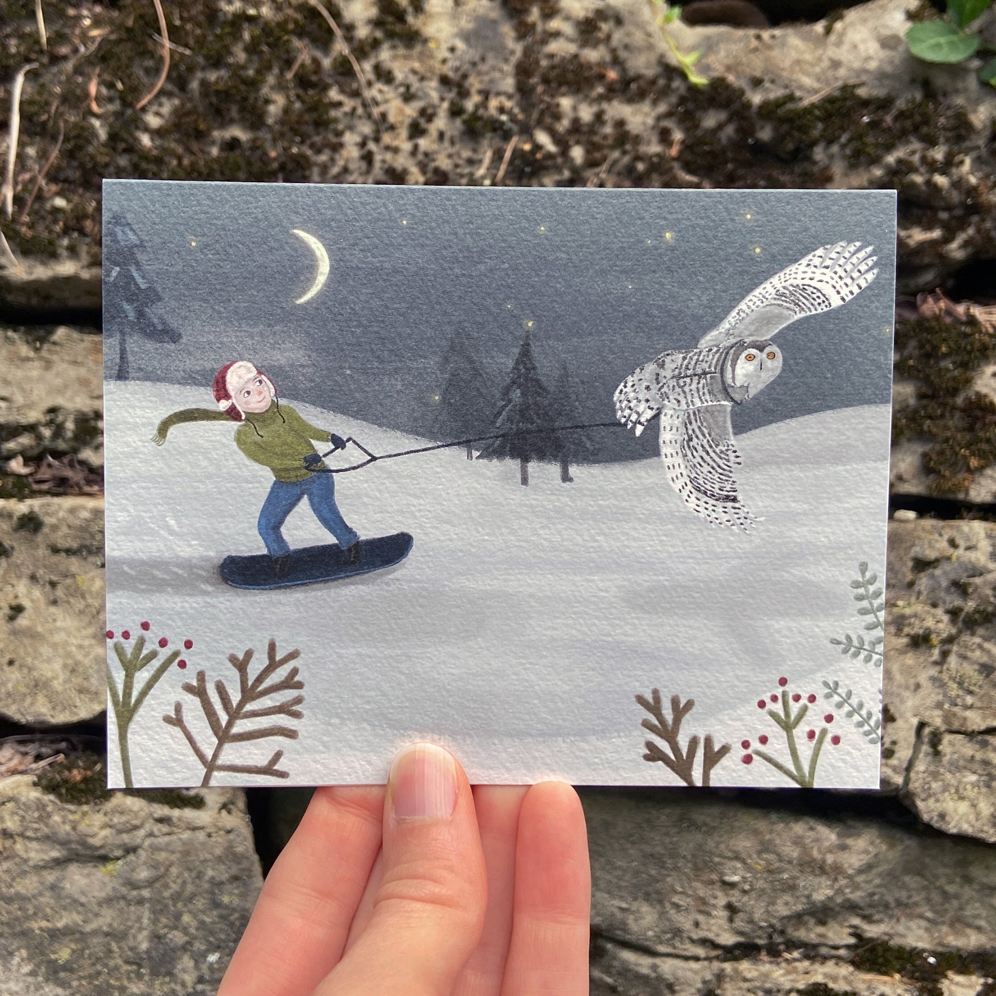Illustrated card with a person snowboarding and an owl, held over a natural background.