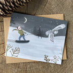 Unique greeting card with a winter scene of a child snowboarding with an owl, on a wooden surface.