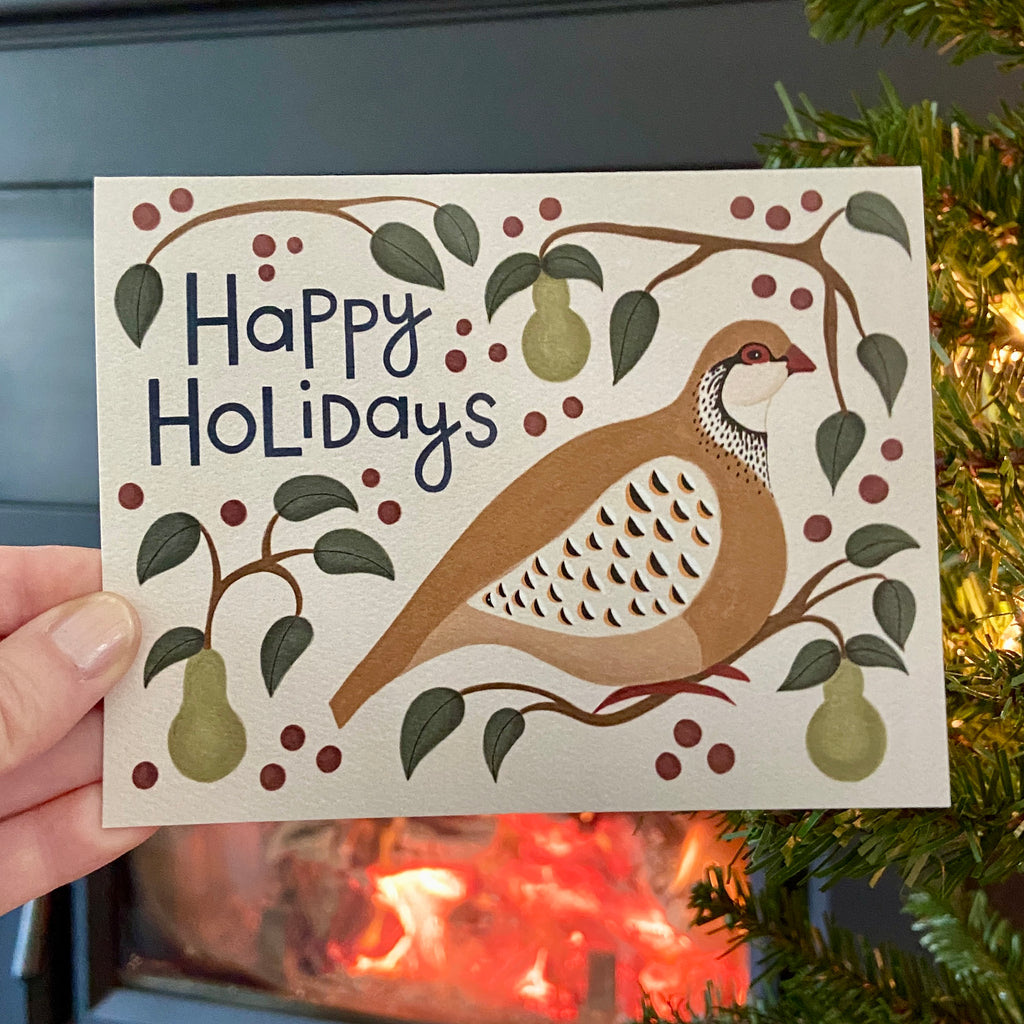 Christmas card with a partridge illustration and 'Happy Holidays' text held in front of a fireplace.