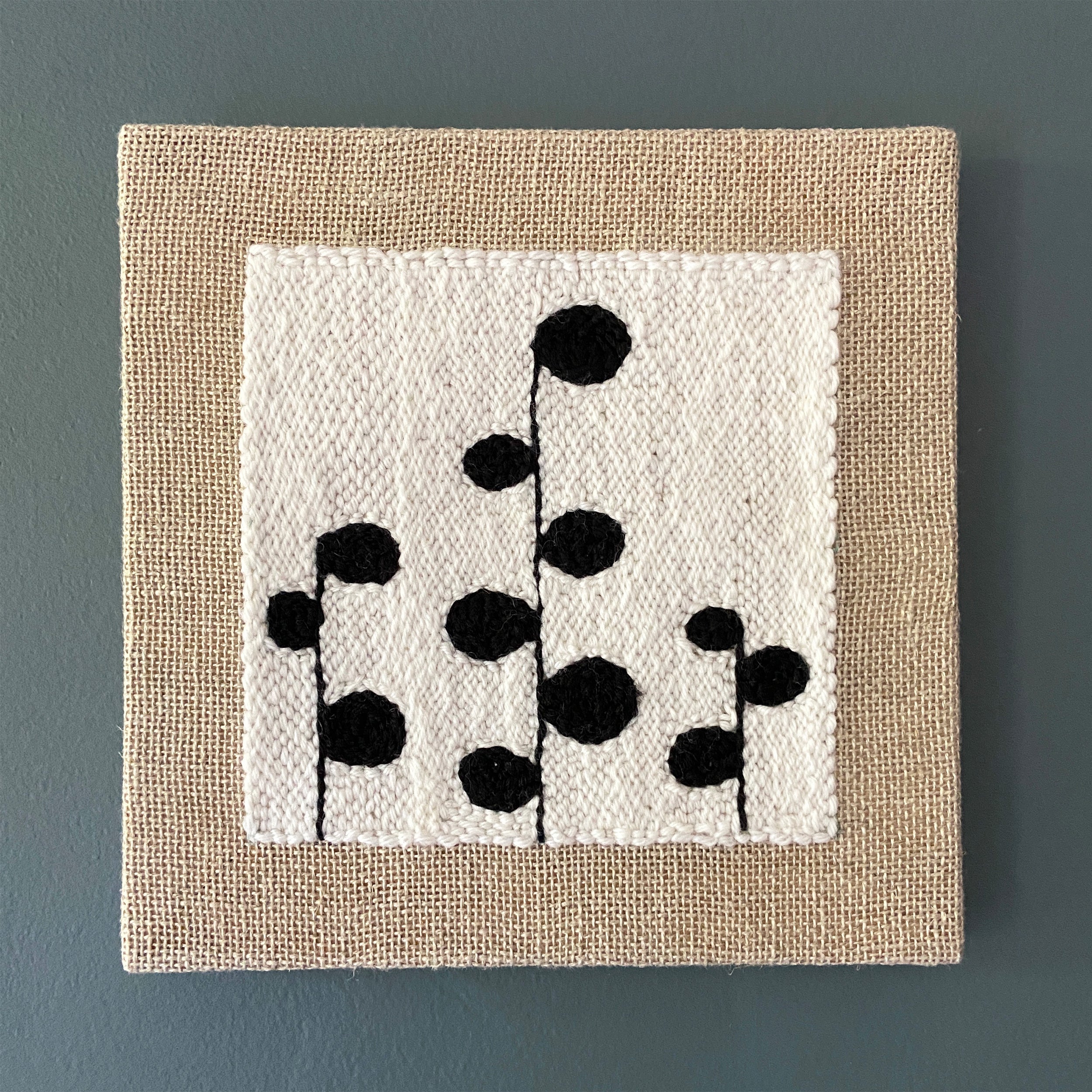 Square wall art with black floral pattern on a beige border against a blue wall