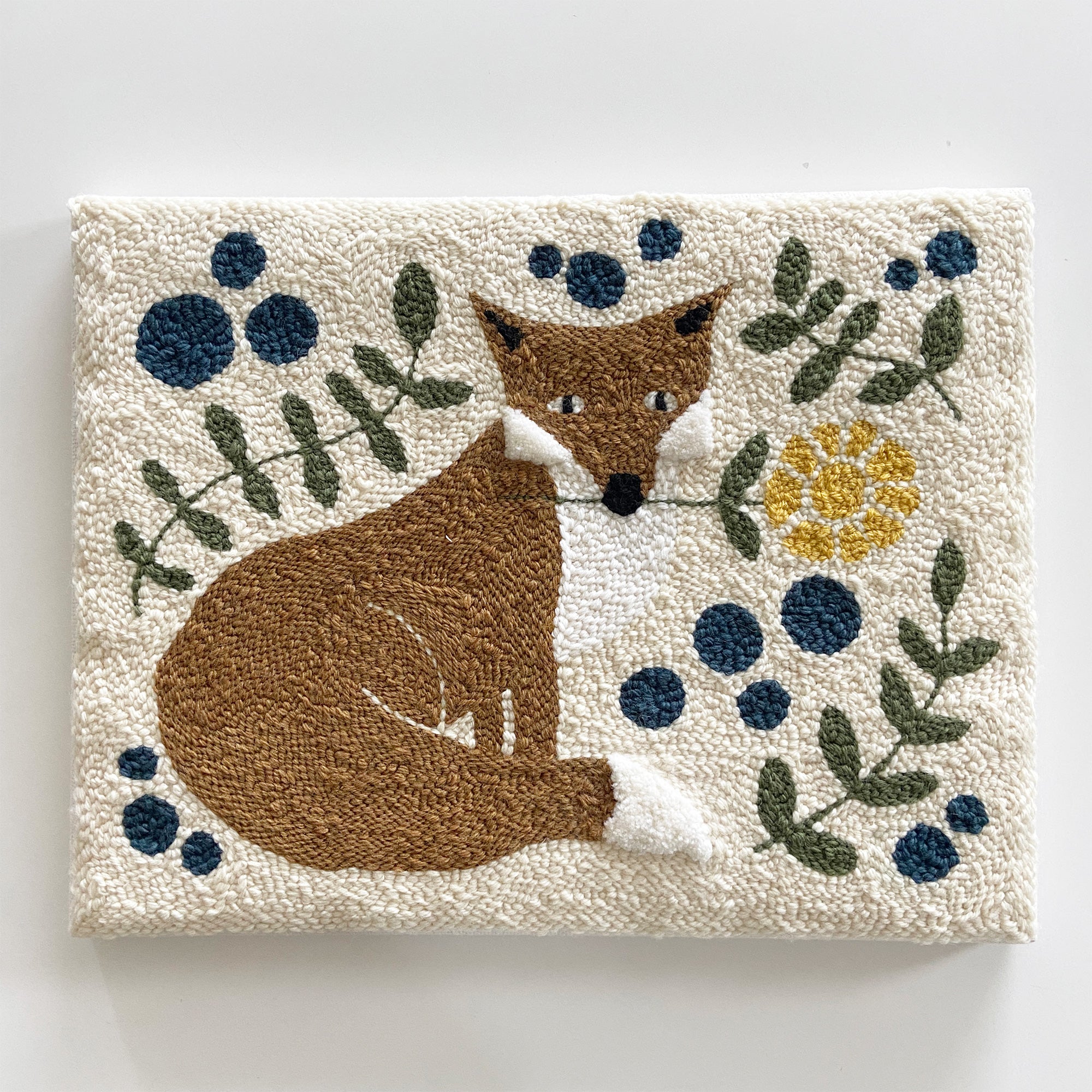 Folk style textile wall art with a red fox and floral elements on a cream background. The fox is holding a yellow flower in its mouth.