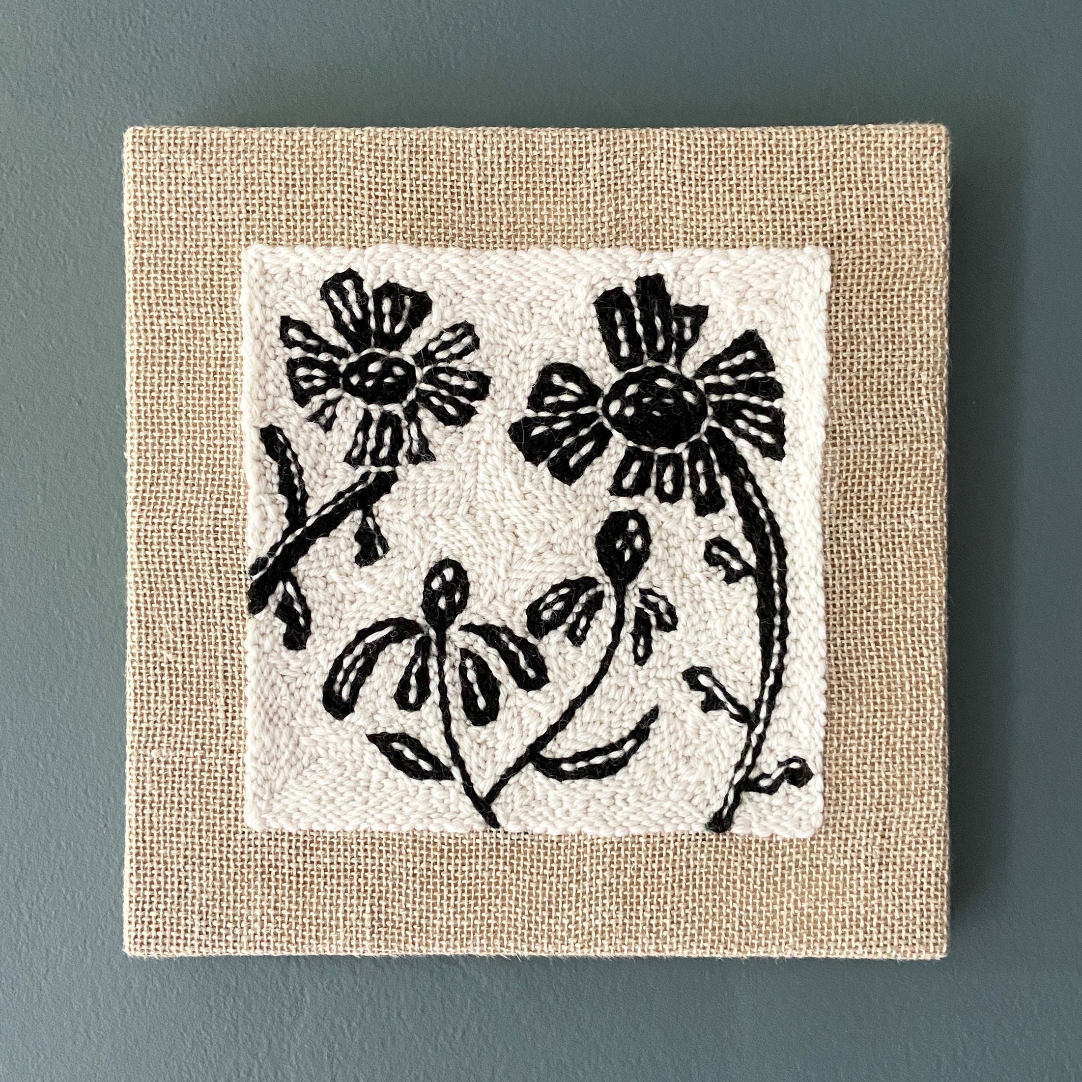 Square wall art with black floral design on a beige border against a gray background