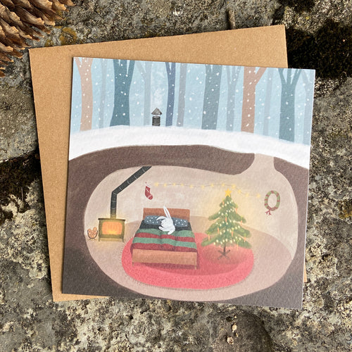 Holiday greeting card featuring a cozy scene of a rabbit sleeping in his underground Christmas decorated home for the winter.