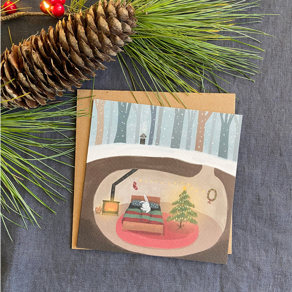 Greeting card with a winter scene of a rabbit's cozy home and Christmas tree, surrounded by pine branches and a pine cone on a textured surface.