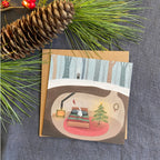 Greeting card with a winter scene of a rabbit's cozy home and Christmas tree, surrounded by pine branches and a pine cone on a textured surface.