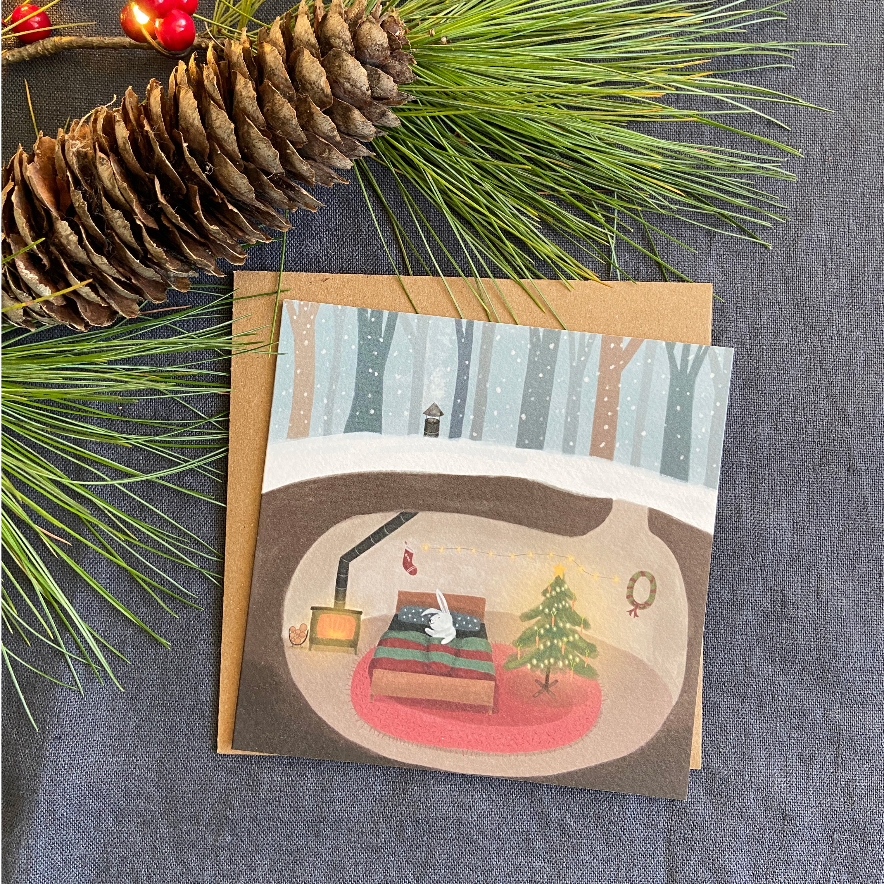 Greeting card with a winter scene of a rabbit's cozy home and Christmas tree, surrounded by pine branches and a pine cone on a textured surface.