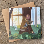 Illustrated greeting card featuring raccoon and rabbit friends in a forest setting, on a wooden surface