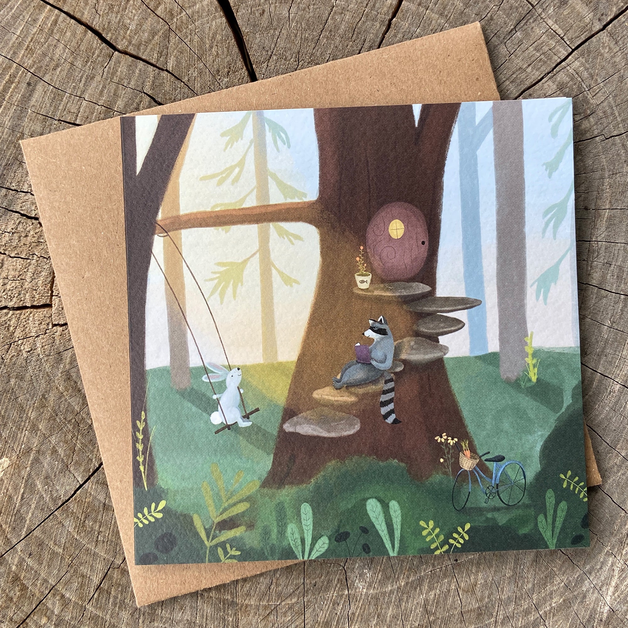 Illustrated greeting card featuring raccoon and rabbit friends in a forest setting, on a wooden surface