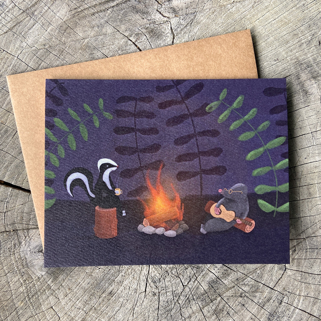 A greeting card featuring a whimsical illustration of a skunk and mole by a campfire, placed on top of a brown kraft envelope over a weathered wood surface.
