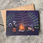 A greeting card featuring a whimsical illustration of a skunk and mole by a campfire, placed on top of a brown kraft envelope over a weathered wood surface.