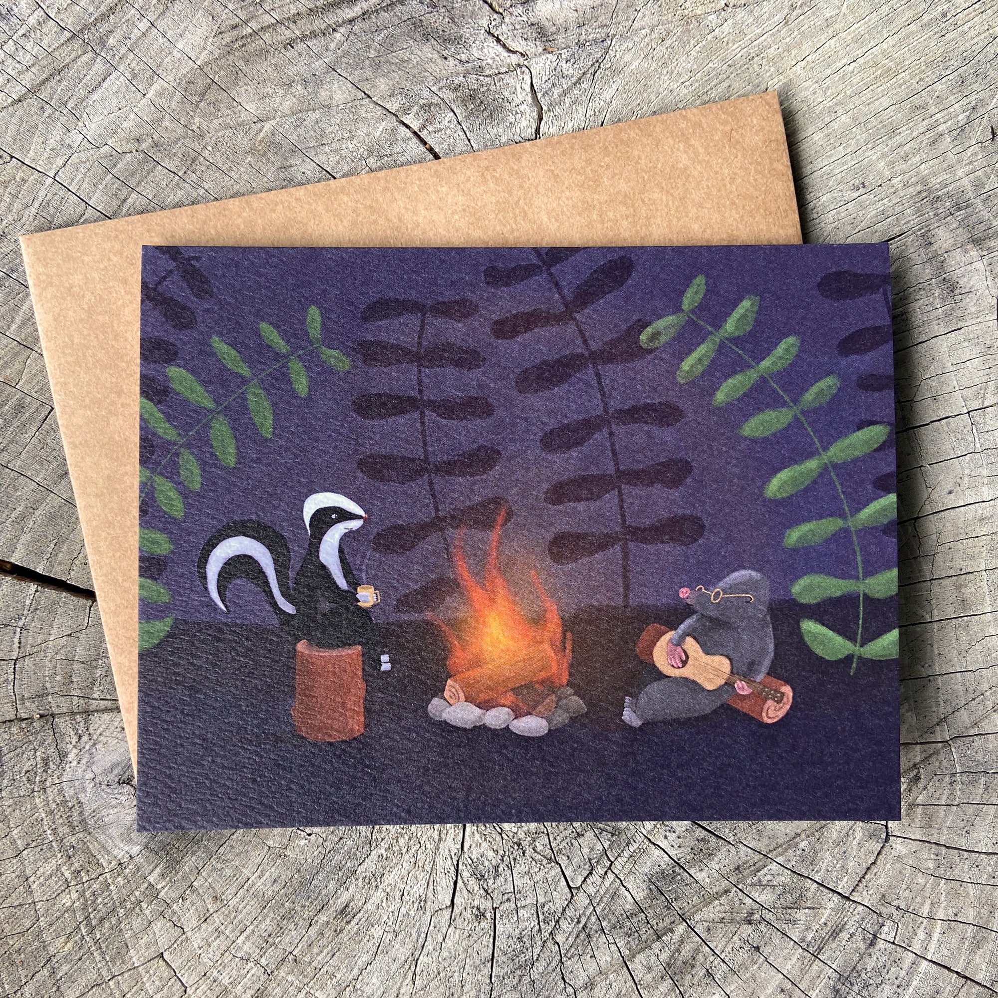 A greeting card featuring a whimsical illustration of a skunk and mole by a campfire, placed on top of a brown kraft envelope over a weathered wood surface.